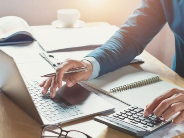 Person typing on a laptop while using a calculator and taking notes at a notebook on a sunny desk.