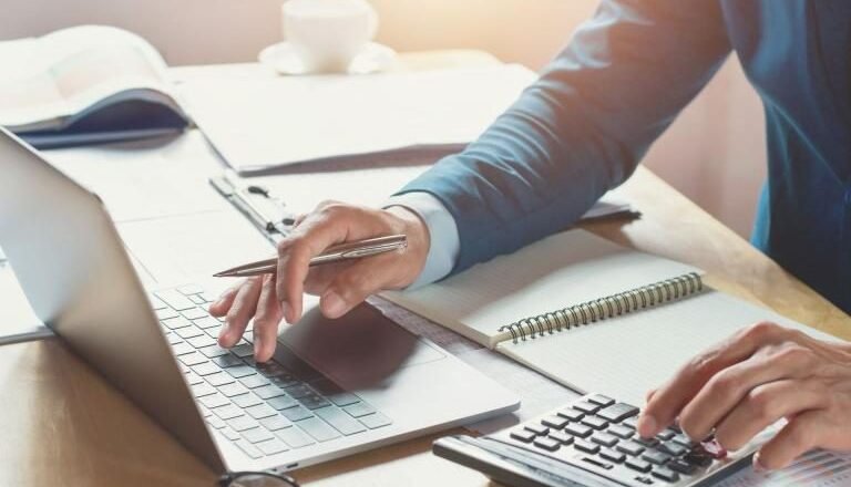 Person typing on a laptop while using a calculator and taking notes at a notebook on a sunny desk.