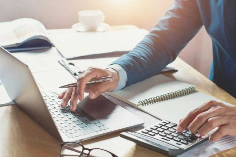 Person typing on a laptop while using a calculator and taking notes at a notebook on a sunny desk.