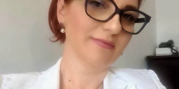 Portrait of a smiling woman with short red hair, black-frame glasses, pearl earrings, and a white blazer in an indoor setting.