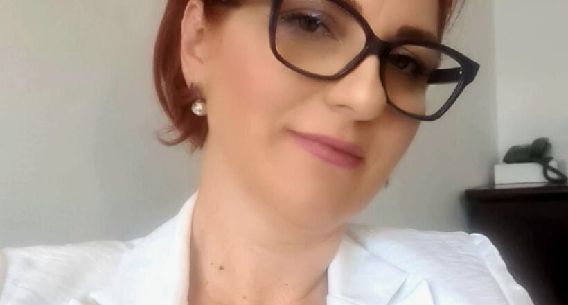 Portrait of a smiling woman with short red hair, black-frame glasses, pearl earrings, and a white blazer in an indoor setting.