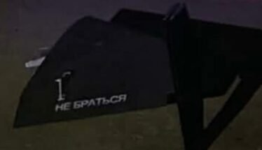 Dark angled object on dirt ground with white stencil text: '1. НЕ БРАТЬСЯ' (Do not touch).