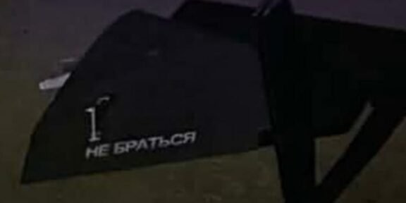 Dark angled object on dirt ground with white stencil text: '1. НЕ БРАТЬСЯ' (Do not touch).