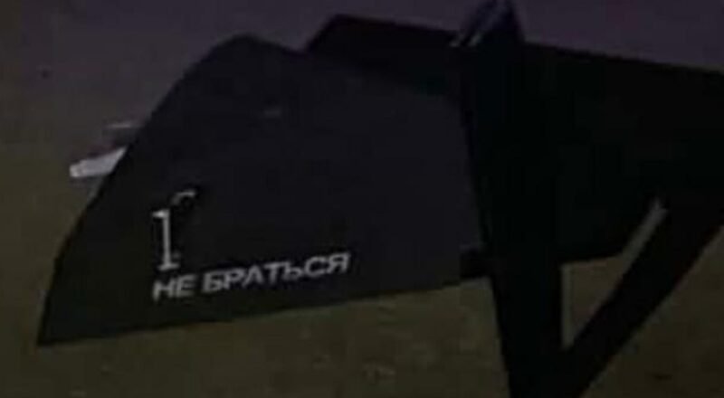 Dark angled object on dirt ground with white stencil text: '1. НЕ БРАТЬСЯ' (Do not touch).