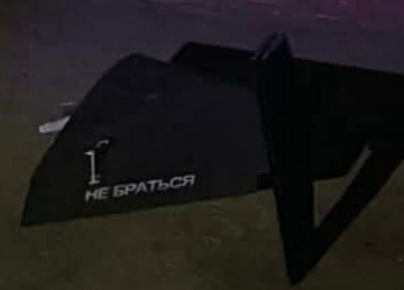 Dark angled object on dirt ground with white stencil text: '1. НЕ БРАТЬСЯ' (Do not touch).