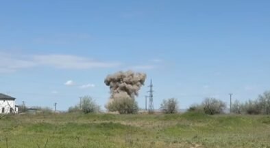 Rural landscape with a large dust plume or explosion near power lines, a house on the left, and a clear blue sky above.