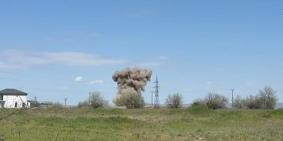 Rural landscape with a large dust plume or explosion near power lines, a house on the left, and a clear blue sky above.