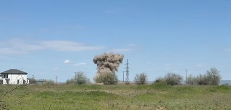Rural landscape with a large dust plume or explosion near power lines, a house on the left, and a clear blue sky above.
