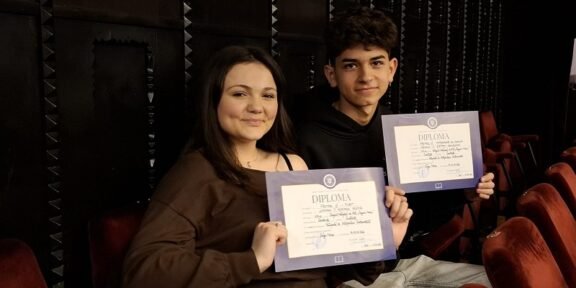 Two students sit in theater seats, smiling at the camera while holding diplomas in purple folders.