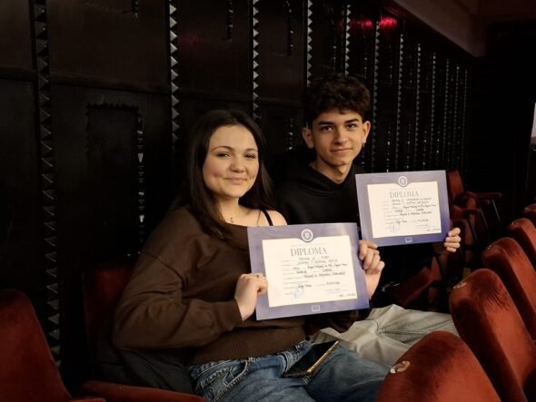 Two students sit in theater seats, smiling at the camera while holding diplomas in purple folders.