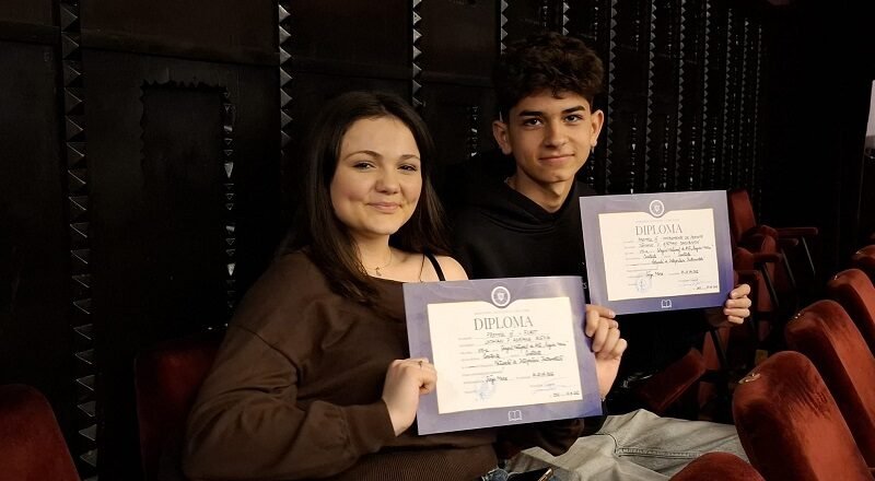 Two students sit in theater seats, smiling at the camera while holding diplomas in purple folders.