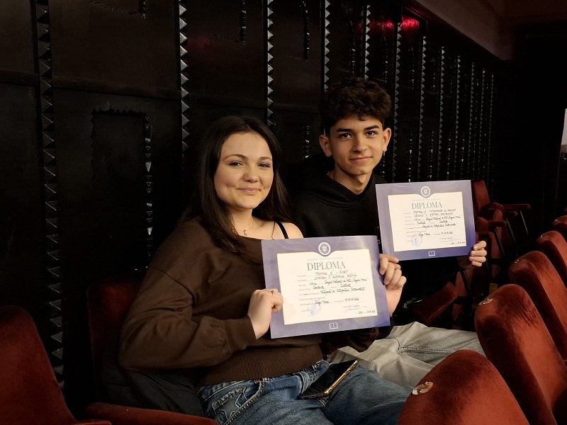 Two students sit in theater seats, smiling at the camera while holding diplomas in purple folders.