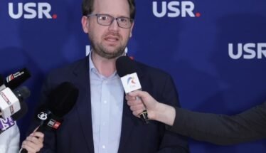 Man in a navy blazer and light blue shirt speaking to reporters, microphones held toward him, against a blue backdrop with USR logos.