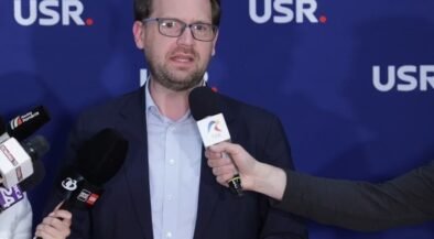 Man in a navy blazer and light blue shirt speaking to reporters, microphones held toward him, against a blue backdrop with USR logos.