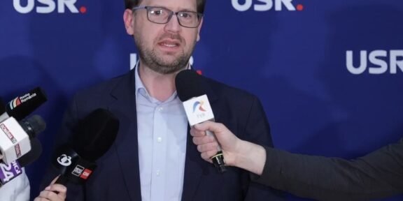 Man in a navy blazer and light blue shirt speaking to reporters, microphones held toward him, against a blue backdrop with USR logos.