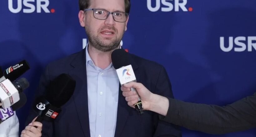 Man in a navy blazer and light blue shirt speaking to reporters, microphones held toward him, against a blue backdrop with USR logos.