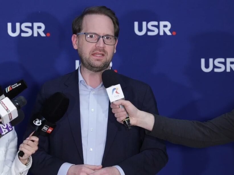 Man in a navy blazer and light blue shirt speaking to reporters, microphones held toward him, against a blue backdrop with USR logos.