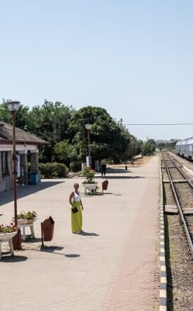 Sunlit train station platform with a stone building on the left and a blue and white passenger train on the tracks to the right.