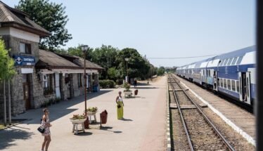 Sunlit train station platform with a stone building on the left and a blue and white passenger train on the tracks to the right.