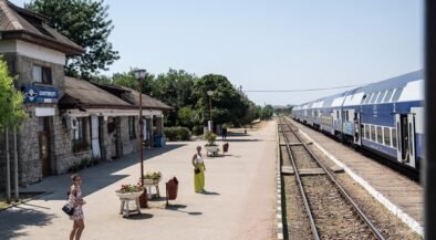 Sunlit train station platform with a stone building on the left and a blue and white passenger train on the tracks to the right.