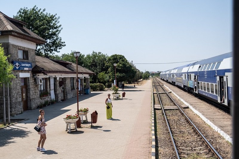 Sunlit train station platform with a stone building on the left and a blue and white passenger train on the tracks to the right.