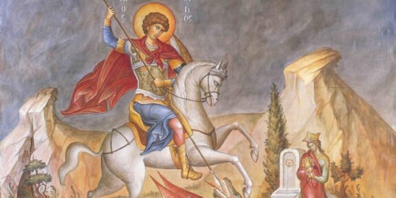 Saint George on a white horse slays a dragon with a spear, red cape billowing, set in a rocky landscape.