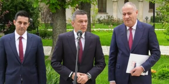 Three suited men stand outdoors at a press conference; center man speaks into a microphone while two colleagues stand by.