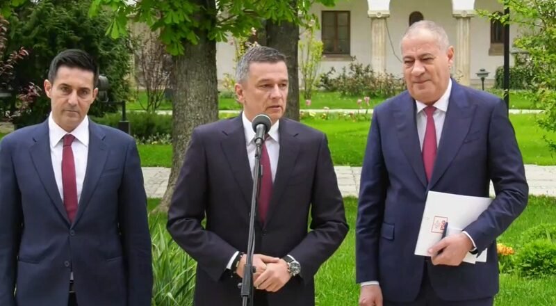 Three suited men stand outdoors at a press conference; center man speaks into a microphone while two colleagues stand by.