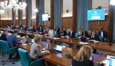Large conference room with many people seated around a long U-shaped table, laptops and microphones in use, chandeliers overhead, presentation screens on walls.