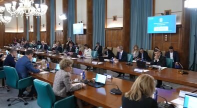 Large conference room with many people seated around a long U-shaped table, laptops and microphones in use, chandeliers overhead, presentation screens on walls.