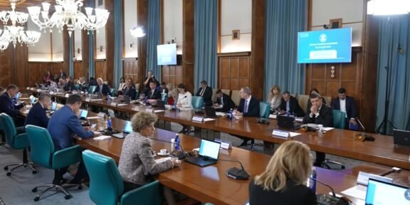Large conference room with many people seated around a long U-shaped table, laptops and microphones in use, chandeliers overhead, presentation screens on walls.