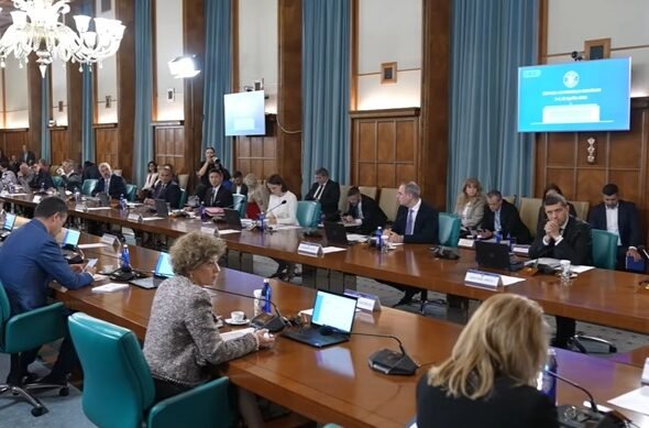 Large conference room with many people seated around a long U-shaped table, laptops and microphones in use, chandeliers overhead, presentation screens on walls.