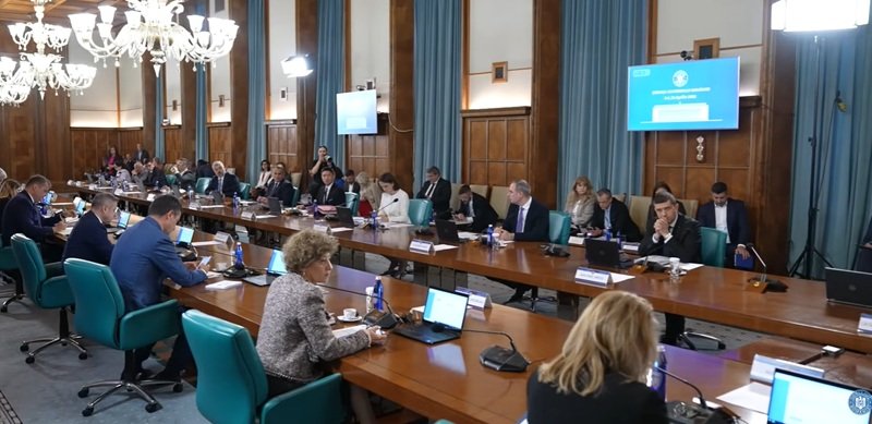 Large conference room with many people seated around a long U-shaped table, laptops and microphones in use, chandeliers overhead, presentation screens on walls.