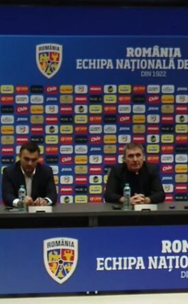 Press conference setup with four men at a long table, Romania football branding on backdrop and podiums in blue. A poster on the left shows a man holding a yellow Romania jersey marked 10 (Hagi).