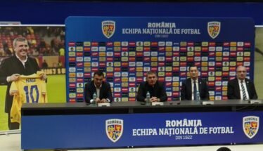Press conference setup with four men at a long table, Romania football branding on backdrop and podiums in blue. A poster on the left shows a man holding a yellow Romania jersey marked 10 (Hagi).