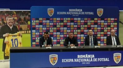 Press conference setup with four men at a long table, Romania football branding on backdrop and podiums in blue. A poster on the left shows a man holding a yellow Romania jersey marked 10 (Hagi).