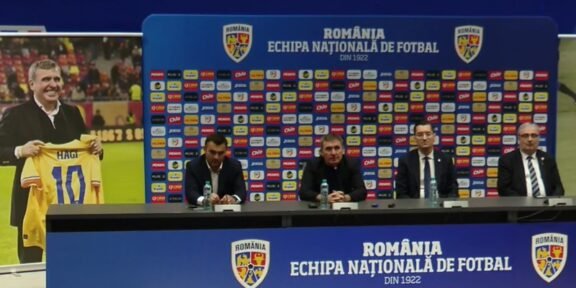 Press conference setup with four men at a long table, Romania football branding on backdrop and podiums in blue. A poster on the left shows a man holding a yellow Romania jersey marked 10 (Hagi).