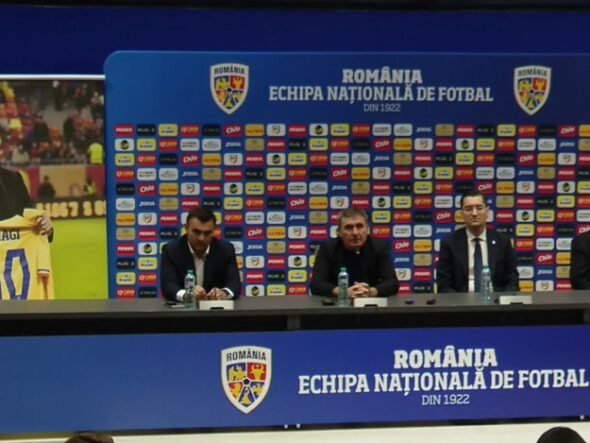 Press conference setup with four men at a long table, Romania football branding on backdrop and podiums in blue. A poster on the left shows a man holding a yellow Romania jersey marked 10 (Hagi).