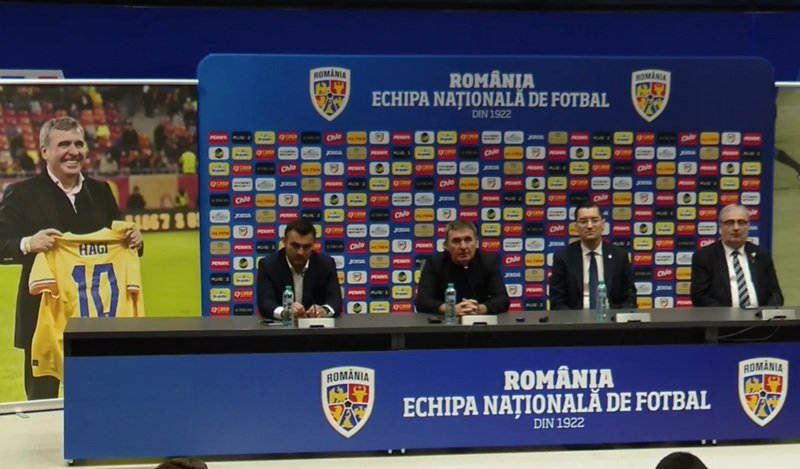 Press conference setup with four men at a long table, Romania football branding on backdrop and podiums in blue. A poster on the left shows a man holding a yellow Romania jersey marked 10 (Hagi).