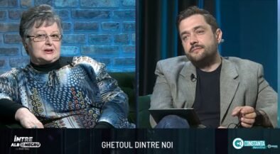 Split-screen interview on a TV talk show: an older woman in a patterned blouse on the left and a man in a gray blazer on the right, seated in a studio with blue brick and dark-blue curtain backgrounds. A bottom caption bar is visible with text.