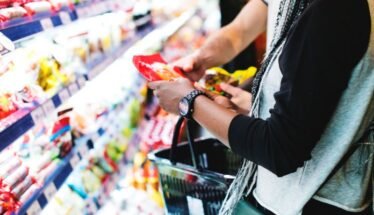 Person selecting a snack from supermarket shelves with a shopping basket nearby