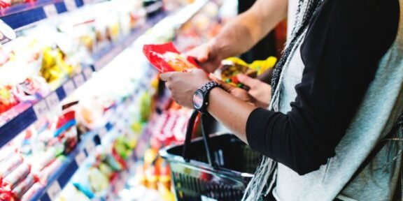 Person selecting a snack from supermarket shelves with a shopping basket nearby