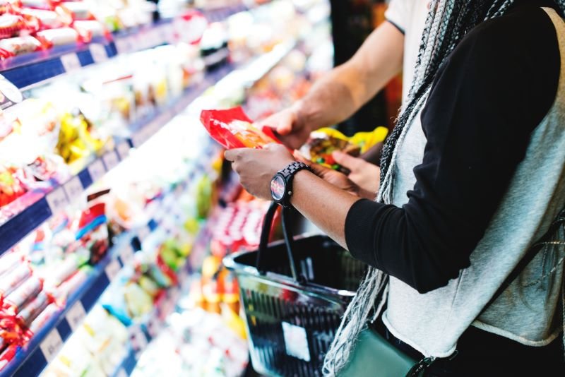 Person selecting a snack from supermarket shelves with a shopping basket nearby