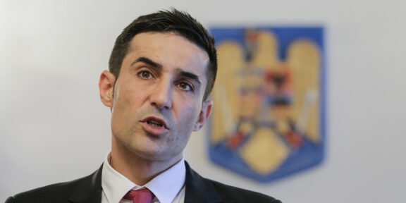 Man in a dark suit and red tie speaking at a press conference, with a blurred emblem in the background.