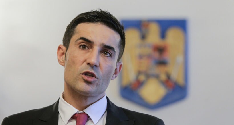 Man in a dark suit and red tie speaking at a press conference, with a blurred emblem in the background.