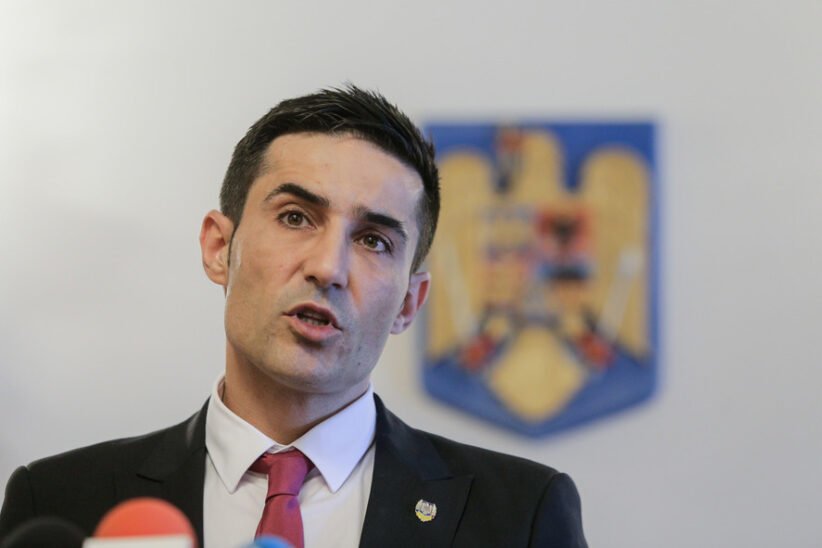 Man in a dark suit and red tie speaking at a press conference, with a blurred emblem in the background.