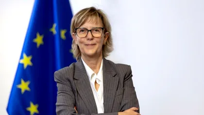 Professional woman in a gray suit standing with arms crossed in front of a European Union flag, conveying a formal, official setting.