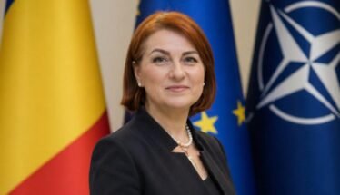 Female official posing in front of European Union and NATO flags, wearing a dark blazer and pearl necklace.