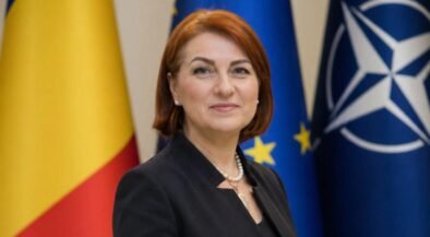 Female official posing in front of European Union and NATO flags, wearing a dark blazer and pearl necklace.