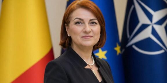 Female official posing in front of European Union and NATO flags, wearing a dark blazer and pearl necklace.